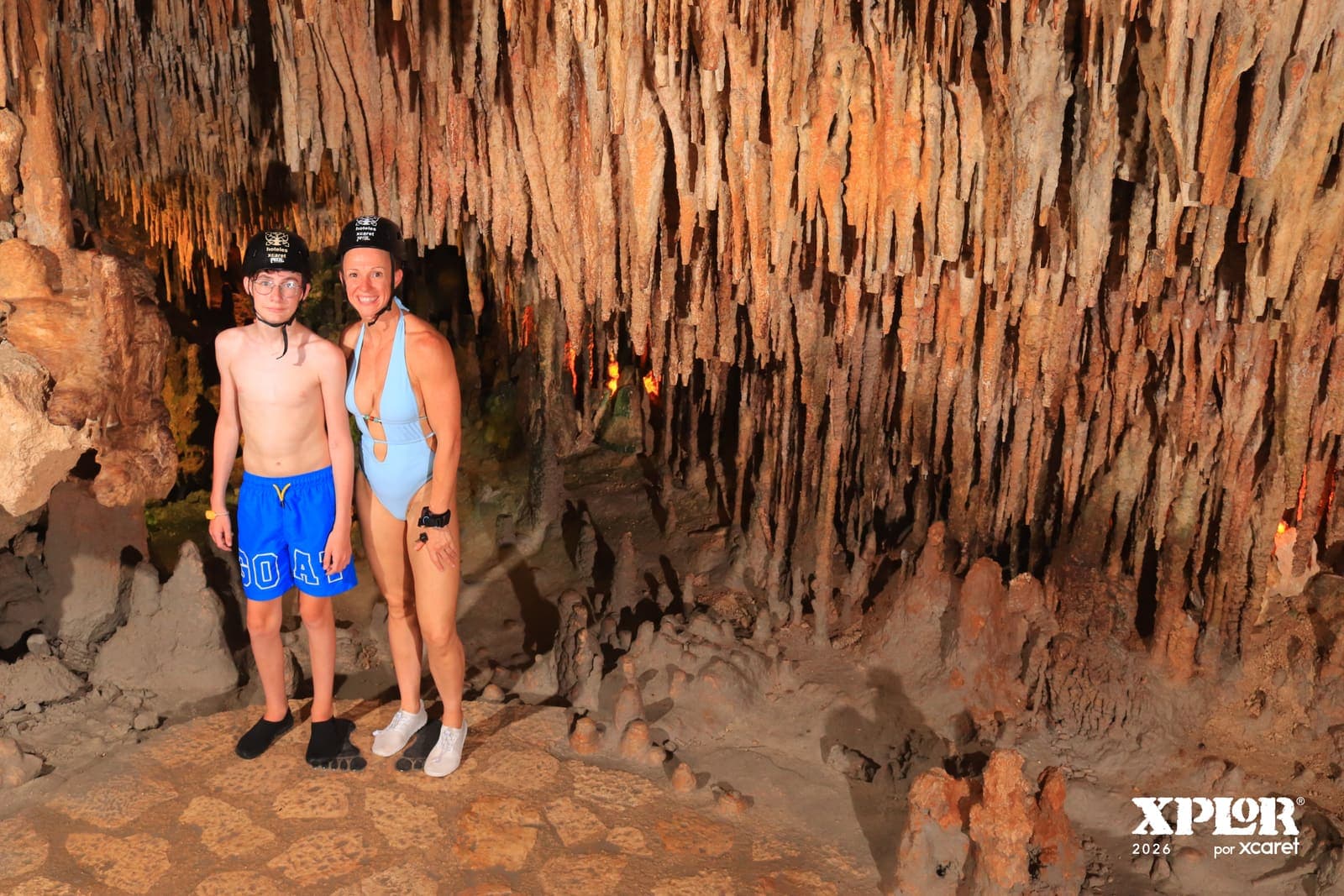 Two visitors in helmets standing beneath dense stalactites in an Xplor cave